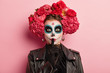 Photo of woman with traditional makeup and flowers on hair, makes hush gesture, keeps index finger over painted lips, prepares for awful death party, dressed in black outfit, isolated on pink