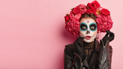 Wall Mural - Studio shot of lovely woman wears halloween makeup, dressed in black outfit, red wreath, has zombie image, looks with scaring expression, isolated over rosy background, free space for your promotion