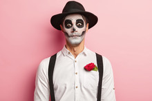 Portrait Of Spooky Handsome Guy Made Makeup For Halloween Event, Has Image Of Vampire Or Ghost, Red Rose Flower In Pocket Of White Shirt, Wears Black Hat, Has Scary Look, Dressed In Zombie Attire