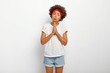Sad begging woman with Afro hair keeps hands in praying gesture, purses lips and asks for huge favour, helping hand in trouble, wears white comfortable t shirt and jean shorts, needs apologise