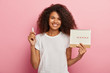Happy curly haired lady holds menstruation calendar with marked pms days and tampon, dressed in casual white t shirt, isolated over pink background, advertises protective absorbent for menses