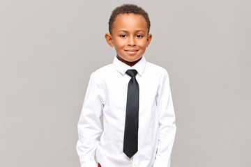 Wall Mural - Waist up studio shot of handsome neat dark skinned male elementary pupil posing isolated dressed in white clean ironed shirt and black elegant tie, ready for going to school, smiling at camera