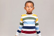 Horizontal shot of confused funny Afro American boy in striped sweater looking away with clueless facial expression, feeling guilty because he ate all sweets, pretending to be innocent. I don't know