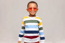 Beauty, Style And Fashion Concept. Picture Of Cheerful Fashionable African Boy Posing Isolated In Studio Wearing Stylish Striped Sweater And Trendy Round Pink Sunglasses, Smiling Happily At Camera