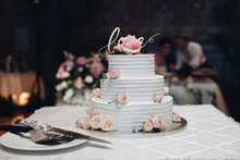 Front View Of Beautiful Wedding Cake With Flowers Staying On Table. Delicious Dessert For Celebration. Lovely Pair At Background. Concept Of Love, Confectionery And Biscuit.