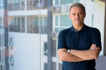 Mature handsome Hispanic man with arms crossed by the glass window at home