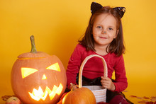 Image Of Little Girl In Cat Mask Sitting And Posing Isolated Over Yelow Background, Child Wearing Red Sweater, Looking Directly At Camera, Holding Trick Or Treat Basket. Halloween Celebration Concept.