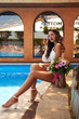 © Dmitry Tsvetkov - Pretty woman with wavy brown hair sitting outdoors near pool with fresh flowers colorful bouquet. Tall slim tanned pretty girl
