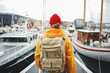 © leonidkos - Man tourist with touristic rucksack wearing yellow jacket walking among authentic fishing boats. Hipster traveler wanderlust with  backpack explore scandinavian city