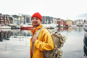 Side view of happy tourist with traveling rucksack standing among fishing boat on city pier. Alone man traveler wearing yellow jacket explore authentic nord town while journey