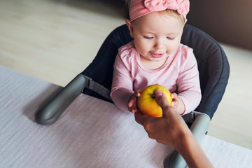 Wall Mural - Nine-month-old smiling baby girl in pink bandage sits at table in highchair and touches yellow apple in her mother's hand.