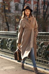 Stylish woman in beige coat and brown hat walking down street in autumn city. Fall fashion look