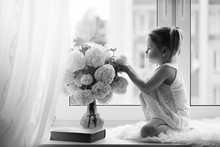 A Little Girl Is Sitting On The Windowsill. A Bouquet Of Flowers In A Vase By The Window And A Girl Sniffing Flowers. A Little Princess In A White Dress With A Bouquet Of White Flowers By Window.