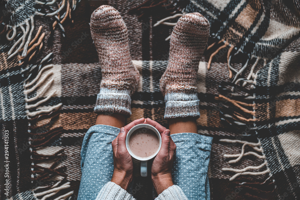 Cozy woman in knitted winter warm socks and in pajamas holding a cup of hot cocoa during resting on checkered plaid blanket at home in winter time. Cozy time and winter drinks. Top view - obrazy, fototapety, plakaty 