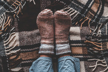 Female Feet In Knitted Winter Warm Socks And In Pajamas On Brown Checkered Plaid Blanket At Home In Cozy Winter Time. Top View