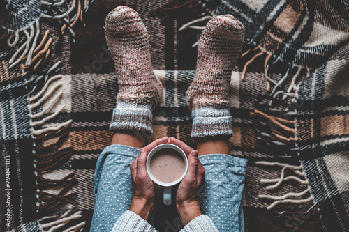 Cozy woman in knitted winter warm socks and in pajamas holding a cup of hot cocoa during resting on checkered plaid blanket at home in winter time Poster Mural XXL