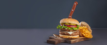 French Fries, Hamburger With Pork Chop And Sauce With Knife Sticking Out Of It On Cutting Board On The Dark Background, Concept Of Fast Food, Long Banner Format
