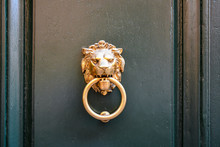 Close-up Of A Golden Metal Door Knocker Representing A Lion's Head Holding A Circle Between The Jaws On A Dark Green Wooden Door, Tuscany, Italy
