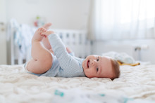 Side View Of Adorable Smiling Caucasian 6 Months Old Baby Boy Dressed In Bodysuit Holding His Feet And Looking At Camera While Lying On Bed In Bedroom.