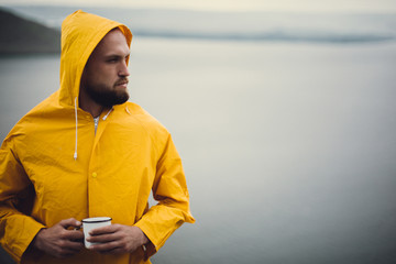 Hipster traveler in yellow raincoat holding metal mug and standing on cliff in rainy windy day with view on lake. Wanderlust and travel. Brutal bearded man hiking. Copy space