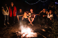 Hipster Man Playing On Acoustic Guitar And Singing Song With Friends Travelers At Big Bonfire At Night Camp In The Forest. Group Of People Chilling At Fire In The Evening, Camping Near Lake