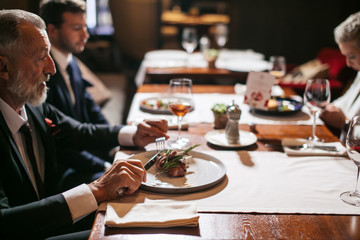 Close up of old bearded man with calm face, holding silver knife and fork ready to start eating delicious meal, in luxury place, horizontal shot