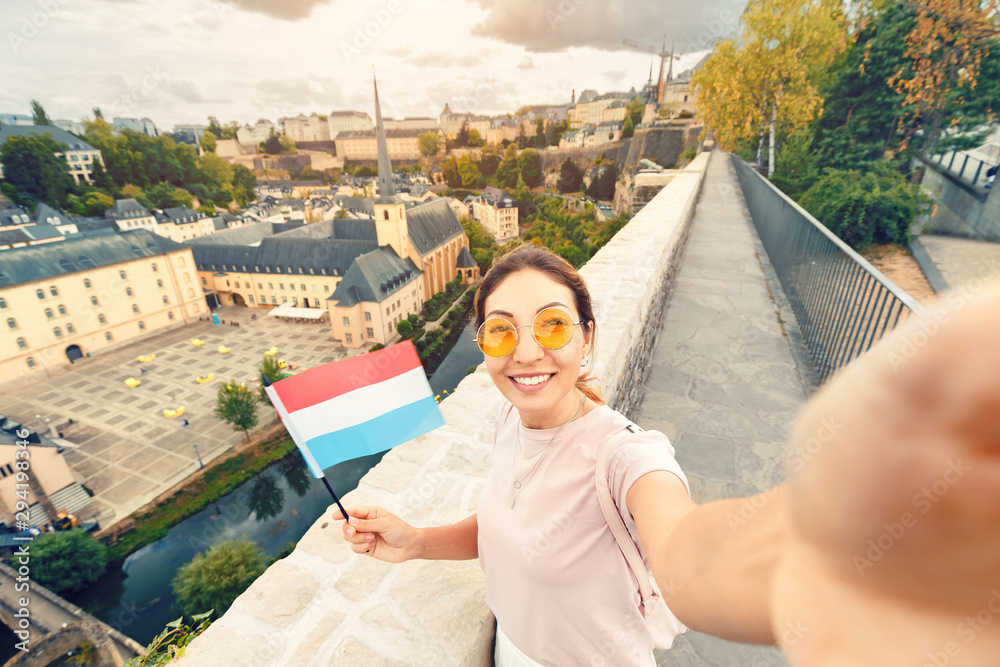 Obraz na płótnie Asian tourist girl takes a selfie with the flag of Luxembourg against the background of the old quarter of the city of Grund and the Church. w salonie