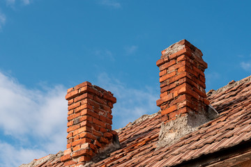 Two traditional red brick chimneys on an old clay tile roof against a blue sky. Chimney in need of sweeping and repair. Preparing for winter concept.