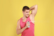 Young man applying deodorant to armpit on yellow background