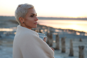 Adult middle-aged blonde woman wrapped herself in a bright plaid on the beach and looks at the sea, selective focus