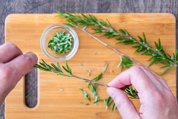 Woman’s hands pulling fresh rosemary leaves off a sprig, sprigs of rosemary with on a bamboo cutting board, chopped rosemary, paring knife and small glass bowl