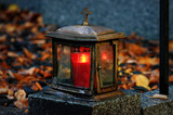 Fototapeta Krajobraz - old damaged grave light made of metal with burning candle on a grave with autumn leaves in blurred background