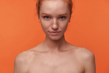 Wall Mural - Studio shot of beautiful young lady with red hair in knot standing over orange background with hands down, smiling softly to camera with raised eyebrow. Human emotion facial expression body language