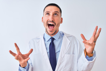 Sticker - Young doctor man wearing white coat over isolated background crazy and mad shouting and yelling with aggressive expression and arms raised. Frustration concept.