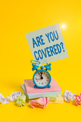 Text sign showing Are You Covered Question. Business photo showcasing asking someone or patient if he have insurance Alarm clock sticky note paper balls stacked notepads colored background