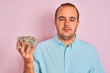 Poster - Young man holding bowl with sunflowers seeds standing over isolated pink background with a confident expression on smart face thinking serious