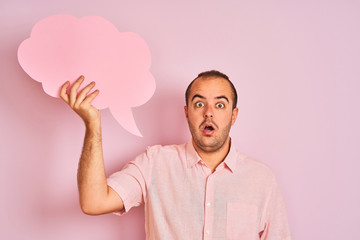 Sticker - Young man holding cloud speech bubble standing over isolated pink background scared in shock with a surprise face, afraid and excited with fear expression