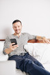 © DragonImages - Smiling cheerful young man sitting on bed when reading funny messages from friends on smartphone