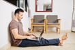 © DragonImages - Barefoot young man sitting on the floor under opened window and programming on laptop