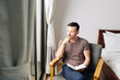 © DragonImages - Pensive young man sitting in armchair in the morning, looking through window and filling gratitude journal