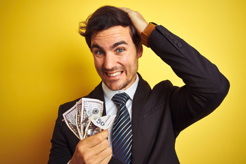 Canvas Print - Young handsome businessman wearing suit holding dollars over isolated yellow background stressed with hand on head, shocked with shame and surprise face, angry and frustrated. Fear