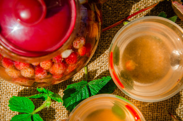 Berry tea. Strawberries in a teapot and a two glass cup with tea with green and red leaves of wild strawberry on a traditional background top view