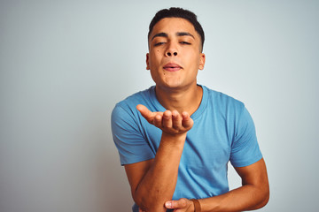 Young brazilian man wearing blue t-shirt standing over isolated white background looking at the camera blowing a kiss with hand on air being lovely and sexy. Love expression.