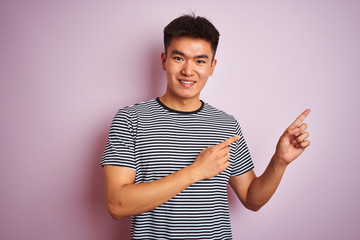 Young asian chinese man wearing striped t-shirt standing over isolated pink background smiling and looking at the camera pointing with two hands and fingers to the side.