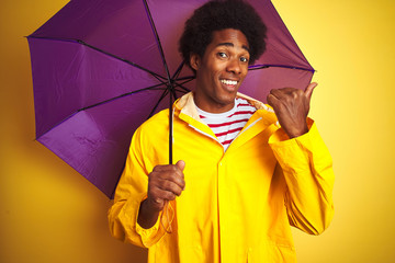 Sticker - Afro american man wearing rain coat and umbrella standing over isolated yellow background pointing and showing with thumb up to the side with happy face smiling