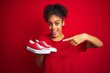 Wall Mural - Young african american woman holding fashion sneakers over isolated red background very happy pointing with hand and finger