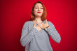 © Krakenimages.com - Young redhead woman wearing strapes navy shirt standing over red isolated background smiling with hands on chest with closed eyes and grateful gesture on face. Health concept.