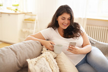 Technology, Communication And Leisure Concept. Pretty Girl With Chubby Cheeks Relaxing At Home With Electronic Gadget. Overweight Young Brunette Woman Texting Sms On Smart Phone And Having Coffee