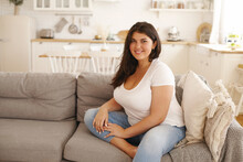 Relaxation, Leisure, Rest, Domesticity And Coziness. Beautiful Charming Young Female With Chubby Cheeks And Curvy Body Sitting On Sofa Barefooted, Keeping One Foot On Floor, Smiling At Camera