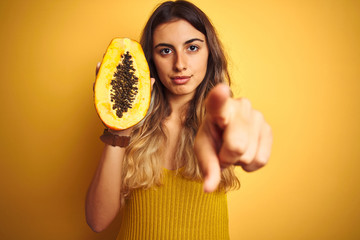 Poster - Young beautiful woman holding papaya over yellow isolated background pointing with finger to the camera and to you, hand sign, positive and confident gesture from the front